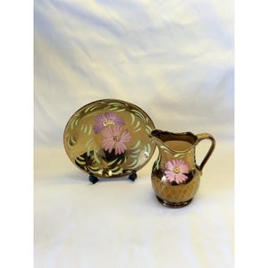Könnte beinhalten: Ein Set aus einem Teller und einem Krug aus metallisch bronzefarbener Keramik. Der Teller ist mit zwei gemalten rosa und violetten Blumen sowie hellgrünen Blattakzenten verziert. Der Krug zeigt eine einzelne rosa Blume und ein Rautenmuster.