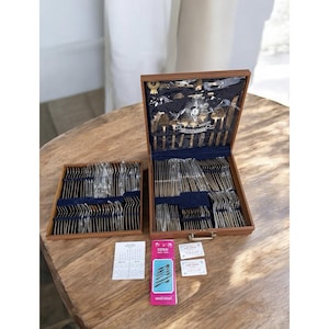 Puede incluir: Un juego de cubiertos vintage en una caja de madera, con una segunda caja más pequeña a la izquierda. El juego incluye tenedores, cuchillos y cucharas, dispuestos cuidadosamente sobre un forro de terciopelo azul. La caja está abierta.