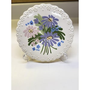 Op de afbeelding: Een rond wit keramieken bord met een geschulpte, reliëf rand, voorzien van een geschilderd boeket van paarse en lichtroze madeliefjes met groene bladeren en kleine blauwe accentbloemen, geplaatst op een doorzichtige standaard.