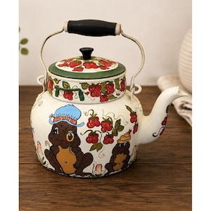 May include: A white metal tea kettle featuring hand-painted illustrations of two brown frogs, one wearing a blue hat, surrounded by red berry clusters and green leaves. The lid has a green border with red berries and a black knob, with a black handle.