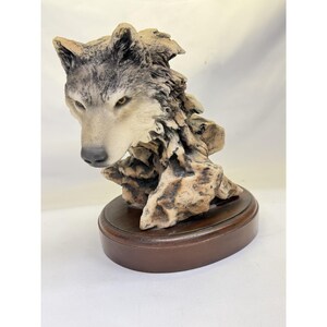 May include: A detailed sculpture of a wolf's head and upper body, with a realistic depiction of fur and facial features. The wolf is mounted on a dark brown, oval wooden base. The sculpture's colors include shades of gray, beige, and brown.
