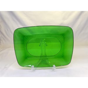 May include: A rectangular, bright green glass serving dish with a black trim. The dish has an oval, ribbed interior and is displayed on a clear, plastic stand. The dish is a vibrant, eye-catching piece.