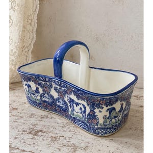 May include: A rectangular ceramic basket with a central arched handle, featuring a cream-coloured interior and a blue rim. The exterior is decorated with a blue, white, and pink pattern depicting farm animals, including cows and roosters.
