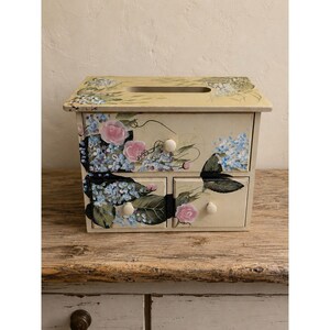 May include: A rectangular wooden tissue box cover with a cream-colored base, featuring hand-painted floral designs of pink roses and blue hydrangea clusters. The unit includes one large top drawer and two smaller bottom drawers, each with a small round knob.