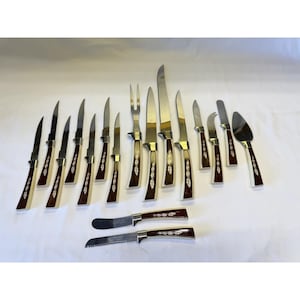 May include: A vintage cutlery set featuring stainless steel blades and brown handles. The set includes various knives, a carving fork, and a cake server. The handles have a decorative inlay design. The knives are arranged on a white surface.