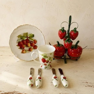 May include: A ceramic set featuring a plate with a strawberry illustration, a mug with strawberry motifs, two lidded strawberry-shaped containers on a metal stand, and four spreaders with white handles decorated with small strawberry patterns.