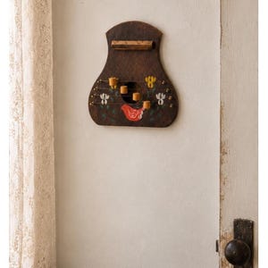 Puede incluir: Un tablero de madera marrón oscuro para montar en la pared con una silueta en forma de campana. Presenta una barra horizontal con pequeños ganchos, un recorte circular central y motivos florales pintados junto a un pájaro rojo.