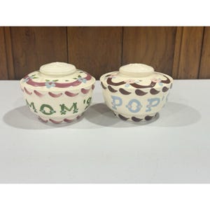 Op de afbeelding: Twee crèmekleurige keramische kommen met bijpassende deksels, elk voorzien van een handgeschilderd bloemenslingermotief in roze, blauw en bruin. De ene kom heeft de groene tekst MOM'S en de andere de blauwe tekst POP'S op de voorzijde.