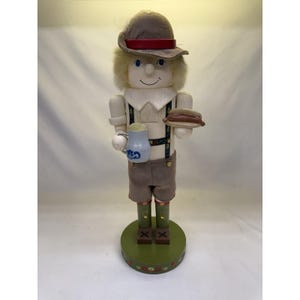 Può includere: Una statuetta schiaccianoci in legno a forma di uomo con un cappello marrone con una fascia rossa, un colletto bianco e pantaloncini marroni. La statuetta tiene in mano una boccale di birra blu e un piatto con un hot dog. La base è verde.