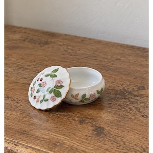 May include: A round white porcelain trinket box with a scalloped edge lid featuring a painted pattern of green leaves, small pink flowers, and red strawberries. The base of the container also displays a matching botanical strawberry motif.