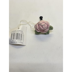 Puede incluir: Un pomo decorativo para muebles con forma de rosa rosa claro con hojas verdes y un poste de metal negro. Una etiqueta de papel blanco está unida por un hilo, con el texto ITEM #: 108420, QUANTITY: 1 EA, MADE IN CHINA y un código de barras.