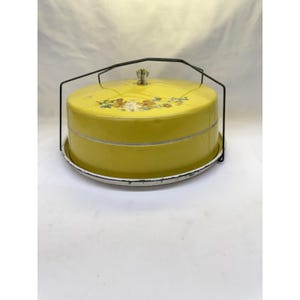 Może przedstawiać: Zabytkowy żółty pojemnik na ciasto z czarnym drucianym uchwytem i kwiatowym wzorem na pokrywie. Pojemnik stoi na białej i żółtej podstawie. Pokrywa ma ozdobny uchwyt w kształcie korony.