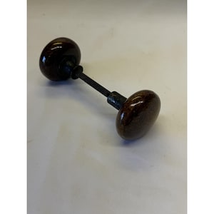 May include: A pair of round, dark brown, glossy door knobs connected by a central metal spindle. The knobs feature a smooth, reflective surface and are attached to a thin, dark gray metal rod that extends between the two circular handles.