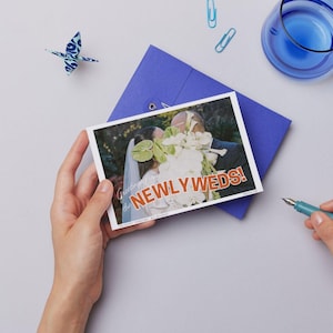May include: A postcard with the text "NEWLY WEDS!" is held over a blue envelope. The postcard features a wedding photo. A blue origami crane, paper clips, and a blue glass are also on the light gray surface.