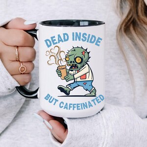 Könnte beinhalten: Weißer Keramikbecher mit schwarzem Innenraum und Griff. Der Becher zeigt eine Cartoon-Zombie-Grafik mit dem Text "DEAD INSIDE BUT CAFFEINATED" in Blau. Der Zombie hält eine Kaffeetasse. Der Becher wird von einer Person gehalten.