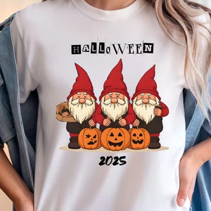以下が含まれることがあります： ハロウィーンのデザインが施された白いTシャツ。赤い帽子をかぶり、カボチャを持った3人のノームが描かれています。「HALLOWEEN」の文字がノームの上に、年号「2025」が下にあります。