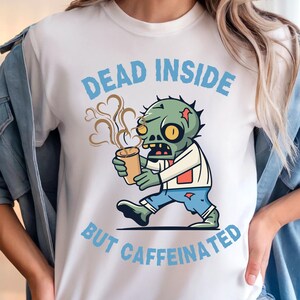 Könnte beinhalten: Weißes T-Shirt mit dem Text "DEAD INSIDE BUT CAFFEINATED" in Hellblau. Das Shirt zeigt einen Cartoon-Zombie, der eine Kaffeetasse hält. Der Zombie ist grün mit gelben Augen und trägt ein weißes Hemd und eine blaue Hose.