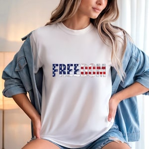 Puede incluir: Camiseta blanca con la palabra "FREEDOM" impresa con los colores de la bandera estadounidense. El diseño es desgastado, con colores rojo, blanco y azul. Estilo casual.