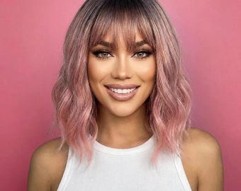May include: A shoulder-length wig featuring wavy, pastel pink hair with darker roots and straight-cut bangs. The hair transitions from a deeper tone at the crown to a lighter, soft pink shade toward the ends, styled with a center part and fringe.