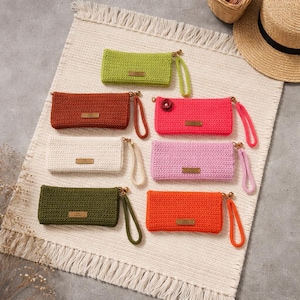 May include: A collection of seven rectangular crochet wristlet pouches in lime green, rust orange, cream, dark olive green, hot pink with a small flower, light pink, and bright orange. Each pouch features a gold-tone metal tag and a matching wrist strap.