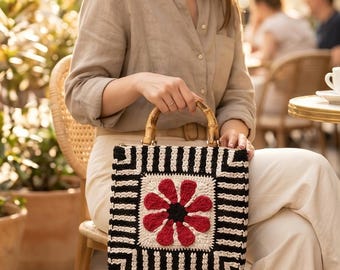 May include: A square crochet handbag featuring a central red flower motif on a cream background, surrounded by a black and white geometric border. The bag includes two curved bamboo handles attached with gold-toned metal hardware.