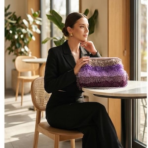May include: A multi-toned, textured clutch in shades of purple, brown, and tan. The bag has a fringed or looped design, and is held by a person wearing a black blazer and pants. The setting appears to be a cafe or restaurant.