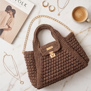 May include: A brown crochet handbag with a gold chain strap and clasp. The bag is next to gold hoop earrings, layered necklaces, and a cup of coffee. A magazine with the word "KINFOLK" is also in the image.