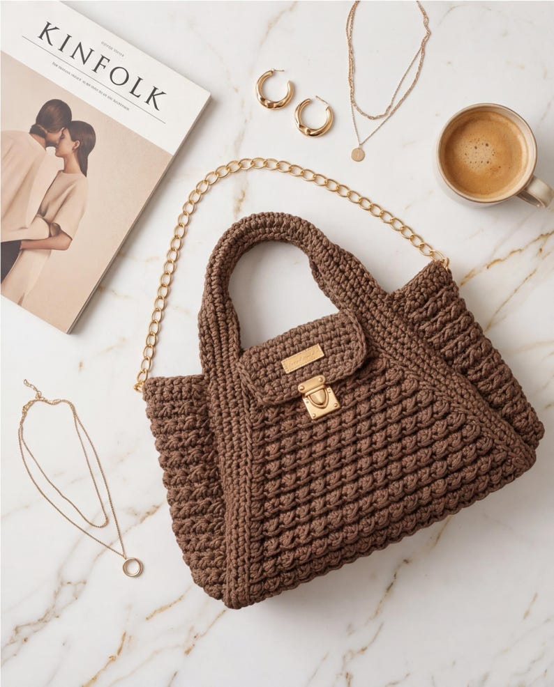May include: A brown crochet handbag with a gold chain strap and clasp. The bag is next to gold hoop earrings, layered necklaces, and a cup of coffee. A magazine with the word "KINFOLK" is also in the image.