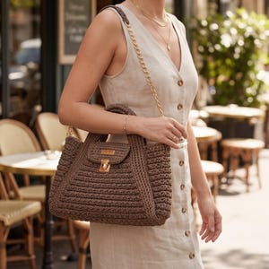 May include: A brown crochet handbag with a flap and gold-tone hardware. The bag has a top handle and a gold chain strap. The bag is being held by a person wearing a beige dress.