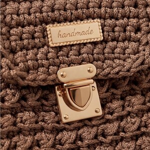 May include: Close-up of a handmade, brown crocheted bag. The bag features a gold-colored clasp and a rectangular gold-colored tag that reads "handmade". The crochet pattern is intricate and detailed, creating a textured surface.