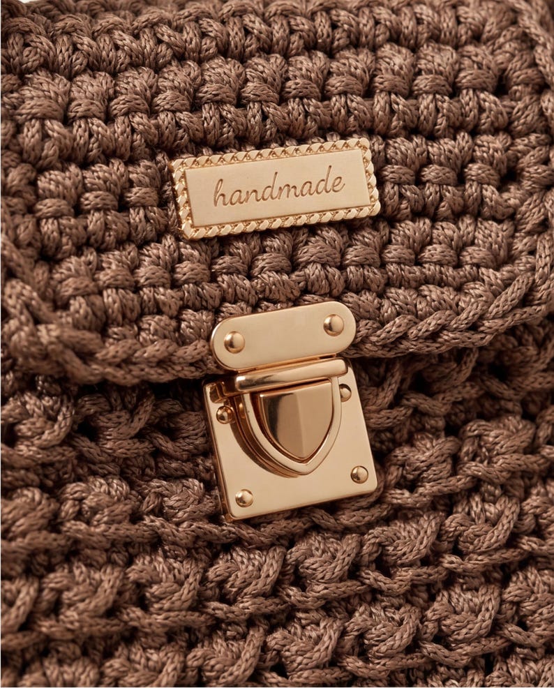 May include: Close-up of a handmade, brown crocheted bag. The bag features a gold-colored clasp and a rectangular gold-colored tag that reads "handmade". The crochet pattern is intricate and detailed, creating a textured surface.