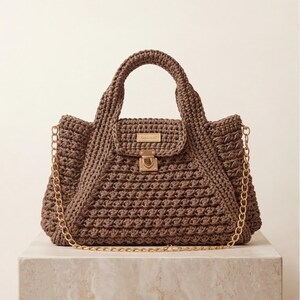 May include: A brown crochet handbag with a top handle and a gold chain strap. The bag features a flap closure with a gold clasp and a small rectangular gold label. The bag is sitting on a marble surface.