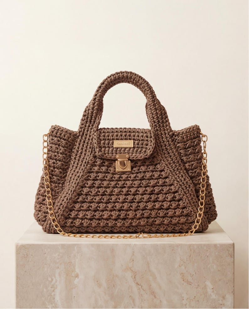 May include: A brown crochet handbag with a top handle and a gold chain strap. The bag features a flap closure with a gold clasp and a small rectangular gold label. The bag is sitting on a marble surface.