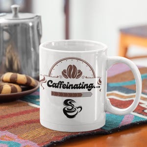 Könnte beinhalten: Weiße Keramik-Kaffeetasse mit dem Text "Caffeinating..." und einem Kaffeebohnen-Design. Die Tasse steht auf einer gemusterten Tischdecke, mit einem Teller mit Keksen und einer Metallkanne im Hintergrund. Eine schwarze Tasse ist ebenfalls sichtbar.