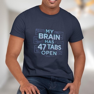 Könnte beinhalten: Marineblaues T-Shirt mit dem Text "MY BRAIN HAS 47 TABS OPEN" in hellblauer, verpixelter Schrift. Das Design zeigt einen Computerbildschirm und Codesymbole. Das T-Shirt hat einen Rundhalsausschnitt und ist aus weichem Material.