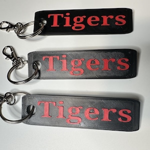 May include: Three rectangular keychains with the word "Tigers" in red lettering. The top keychain is black, while the other two are gray. Each keychain has a silver-colored clasp and ring.