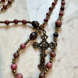May include: A rosary featuring smooth, round pink and dark brown mottled beads interspersed with small, metallic bronze-toned spacer beads. The centerpiece is an ornate, antique-style bronze crucifix with a detailed figure of Jesus and INRI text.