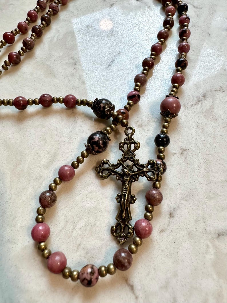 May include: A rosary featuring smooth, round pink and dark brown mottled beads interspersed with small, metallic bronze-toned spacer beads. The centerpiece is an ornate, antique-style bronze crucifix with a detailed figure of Jesus and INRI text.