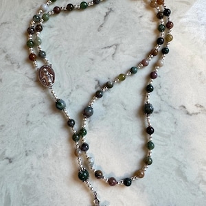 May include: A rosary featuring multi-colored natural stone beads in shades of green, brown, and grey, interspersed with small silver-toned metal spacer beads. The rosary includes a silver-toned metal crucifix and a central oval medal depicting a figure.