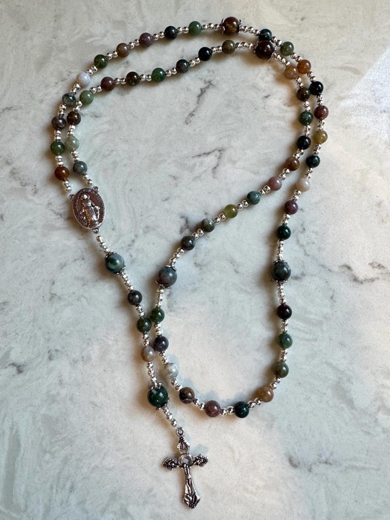 May include: A rosary featuring multi-colored natural stone beads in shades of green, brown, and grey, interspersed with small silver-toned metal spacer beads. The rosary includes a silver-toned metal crucifix and a central oval medal depicting a figure.