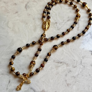 May include: A rosary featuring brown tiger eye gemstone beads interspersed with small gold-toned metal spacer beads. The piece includes a gold-toned metal crucifix and a central oval medal depicting a figure, connected by a gold-toned metal chain.