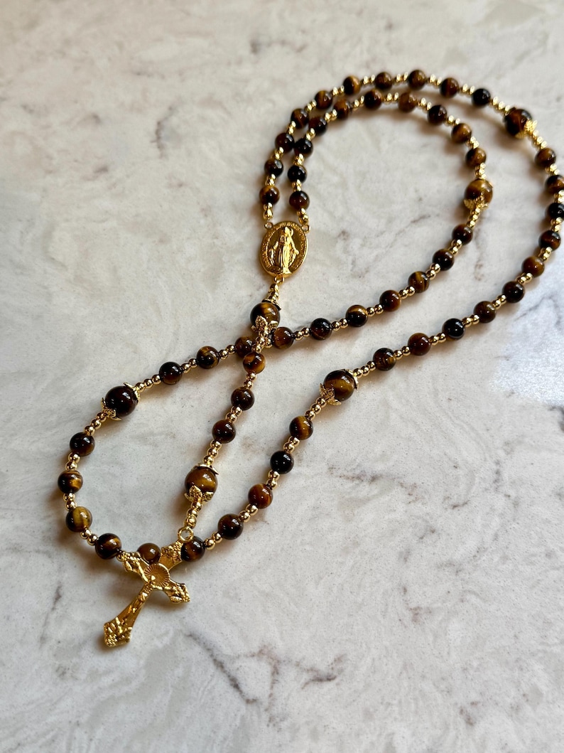 May include: A rosary featuring brown tiger eye gemstone beads interspersed with small gold-toned metal spacer beads. The piece includes a gold-toned metal crucifix and a central oval medal depicting a figure, connected by a gold-toned metal chain.