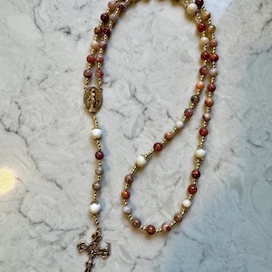 May include: A rosary featuring multi-colored stone beads in shades of red, brown, and cream, interspersed with small gold-toned metal beads. It includes a gold-toned metal crucifix and a central oval medal depicting a religious figure.