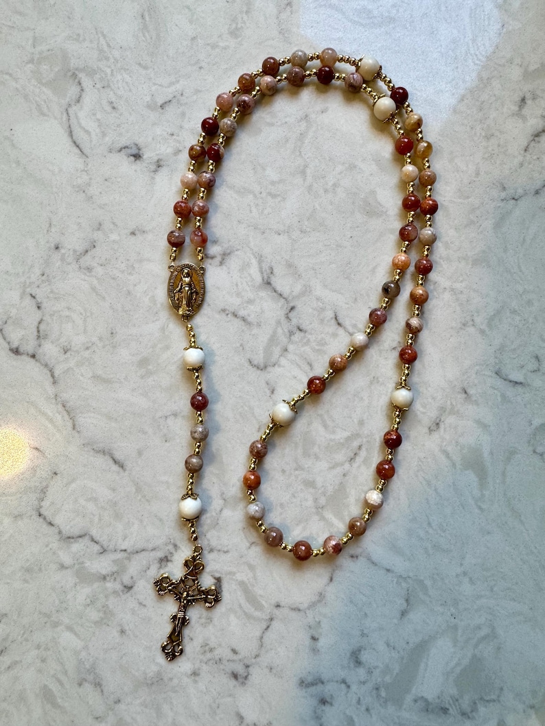 May include: A rosary featuring multi-colored stone beads in shades of red, brown, and cream, interspersed with small gold-toned metal beads. It includes a gold-toned metal crucifix and a central oval medal depicting a religious figure.