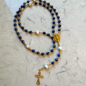 May include: A rosary featuring deep blue lapis lazuli beads interspersed with white marbled beads and small gold-toned spacer beads. The piece includes a gold-toned metal crucifix and a central oval medal depicting a religious figure in relief.