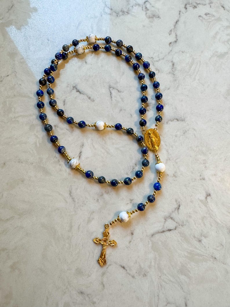 May include: A rosary featuring deep blue lapis lazuli beads interspersed with white marbled beads and small gold-toned spacer beads. The piece includes a gold-toned metal crucifix and a central oval medal depicting a religious figure in relief.