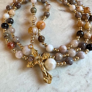 May include: A rosary featuring multi-colored, polished stone beads in shades of brown, cream, gray, and orange, interspersed with small gold-toned metal beads. The rosary includes a gold-toned metal crucifix and a small gold-toned religious medal.
