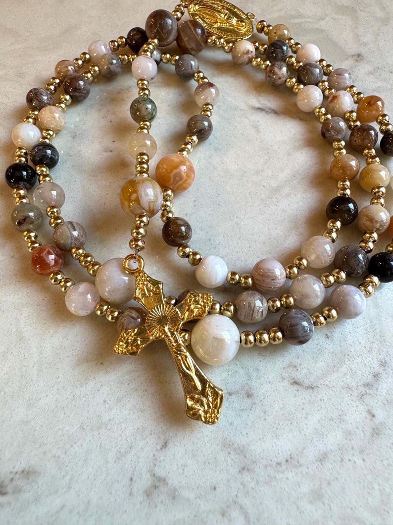 May include: A rosary featuring multi-colored, polished stone beads in shades of brown, cream, gray, and orange, interspersed with small gold-toned metal beads. The rosary includes a gold-toned metal crucifix and a small gold-toned religious medal.