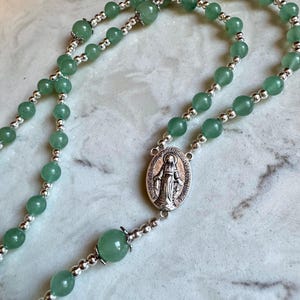 May include: A rosary featuring smooth, round green aventurine beads interspersed with small, polished silver-tone metal spacer beads. The centerpiece is an oval silver-tone medal depicting a figure in robes with raised arms.