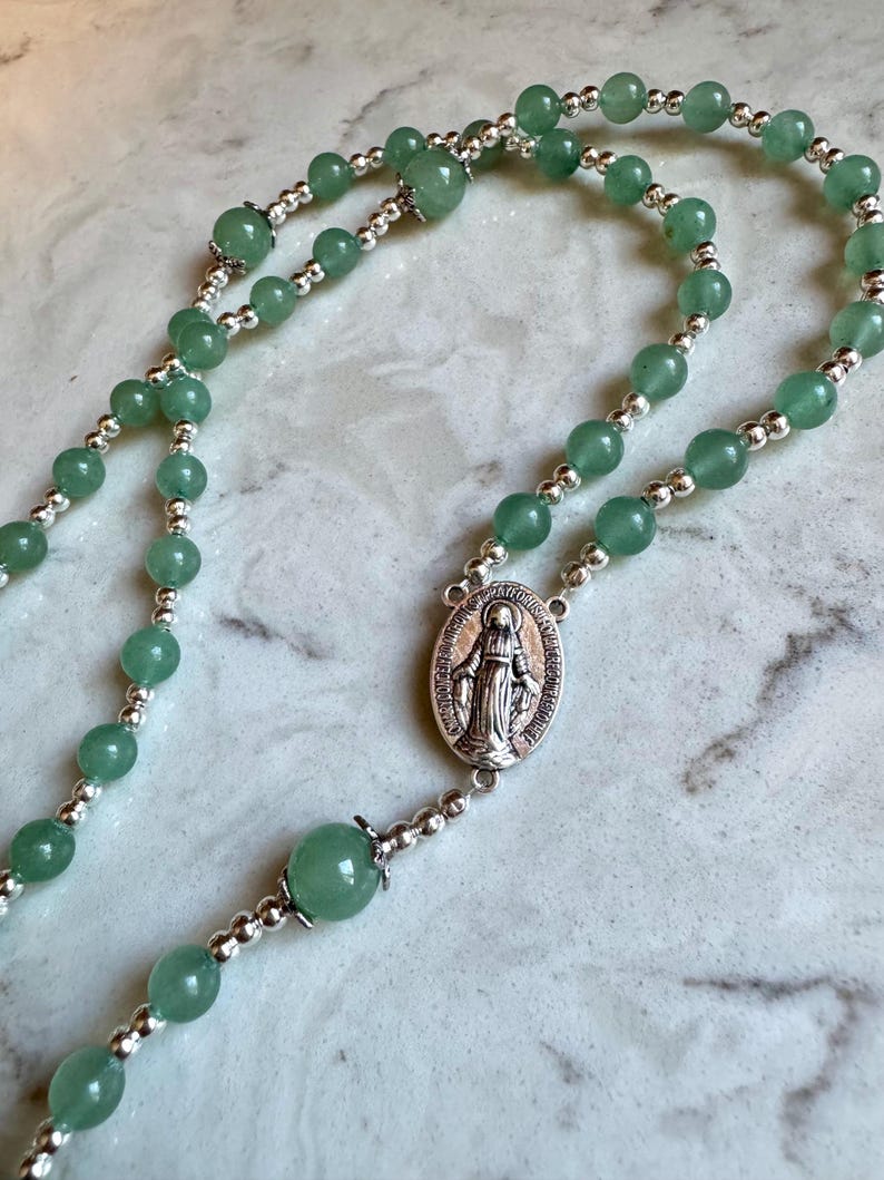 May include: A rosary featuring smooth, round green aventurine beads interspersed with small, polished silver-tone metal spacer beads. The centerpiece is an oval silver-tone medal depicting a figure in robes with raised arms.